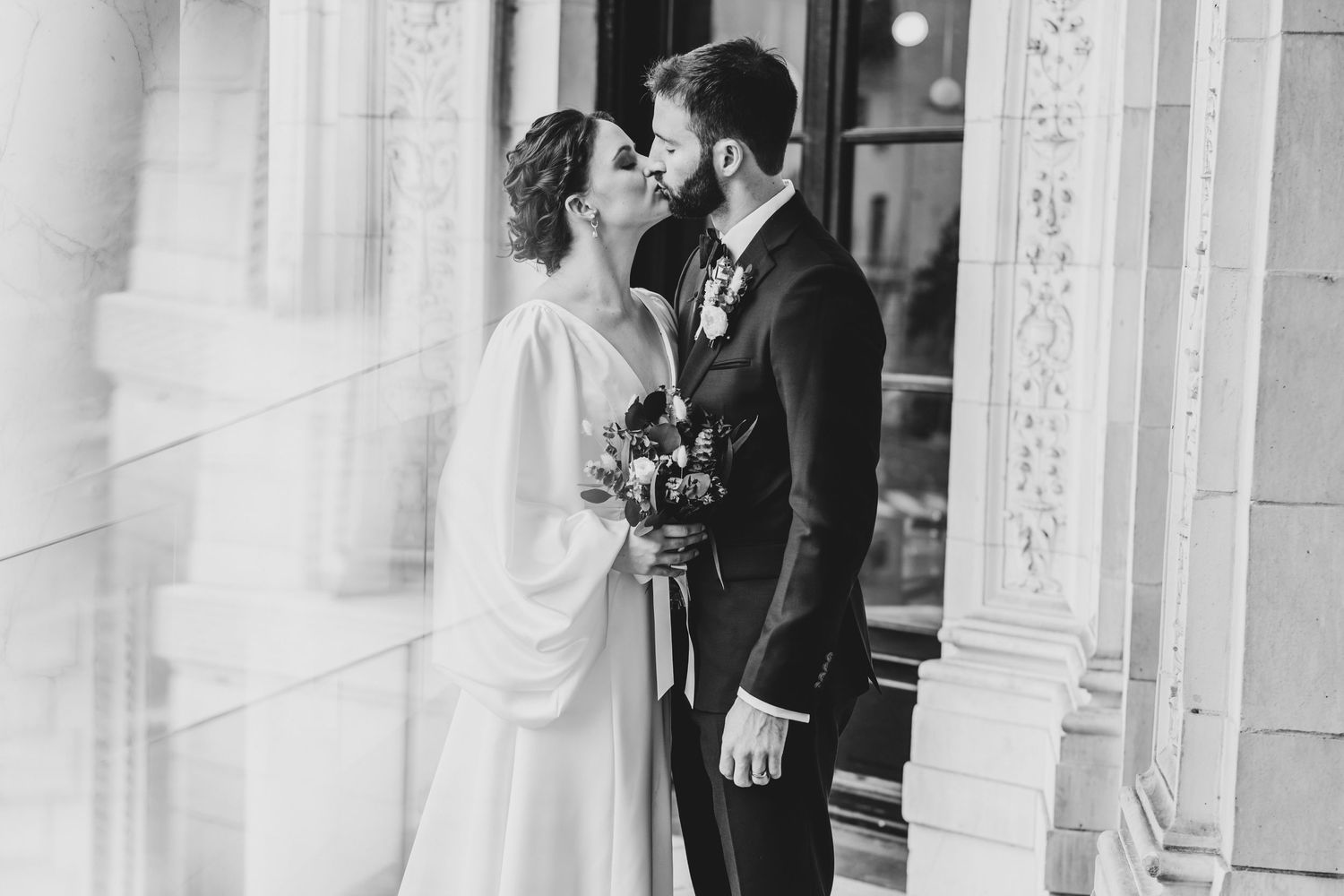 A romantic black and white wedding photo of a couple sharing a tender kiss against an ornate architectural backdrop.