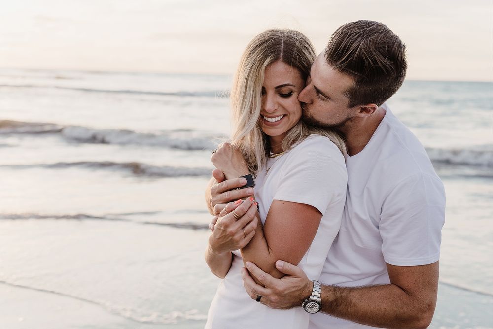 A romantic couple embraces on the beach with the ocean and sunset behind them.