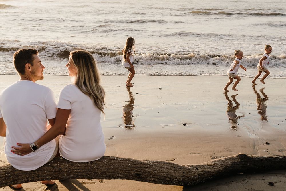 Two people sit on driftwood watching children play at the beach during golden hour.
