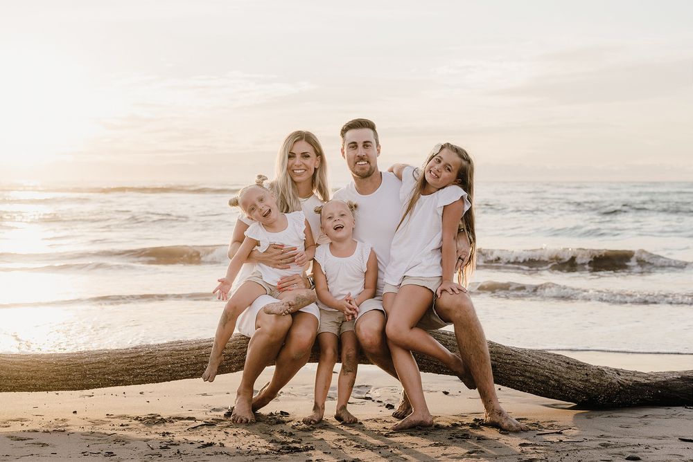 A happy group poses together on a large piece of driftwood at the beach during sunset.
