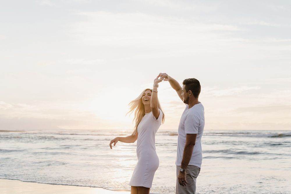 A couple dances together on the beach at sunset with ocean waves in the background.