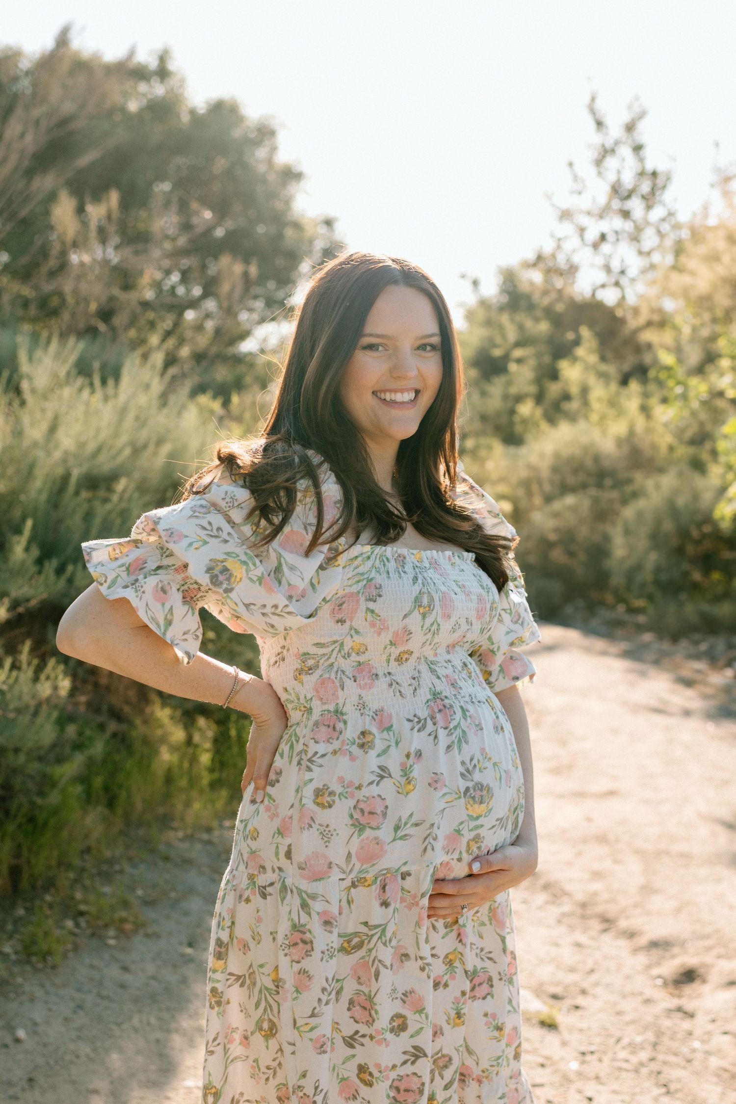 An expectant mother in a floral dress poses on a sunlit nature path during maternity photoshoot in San Diego.