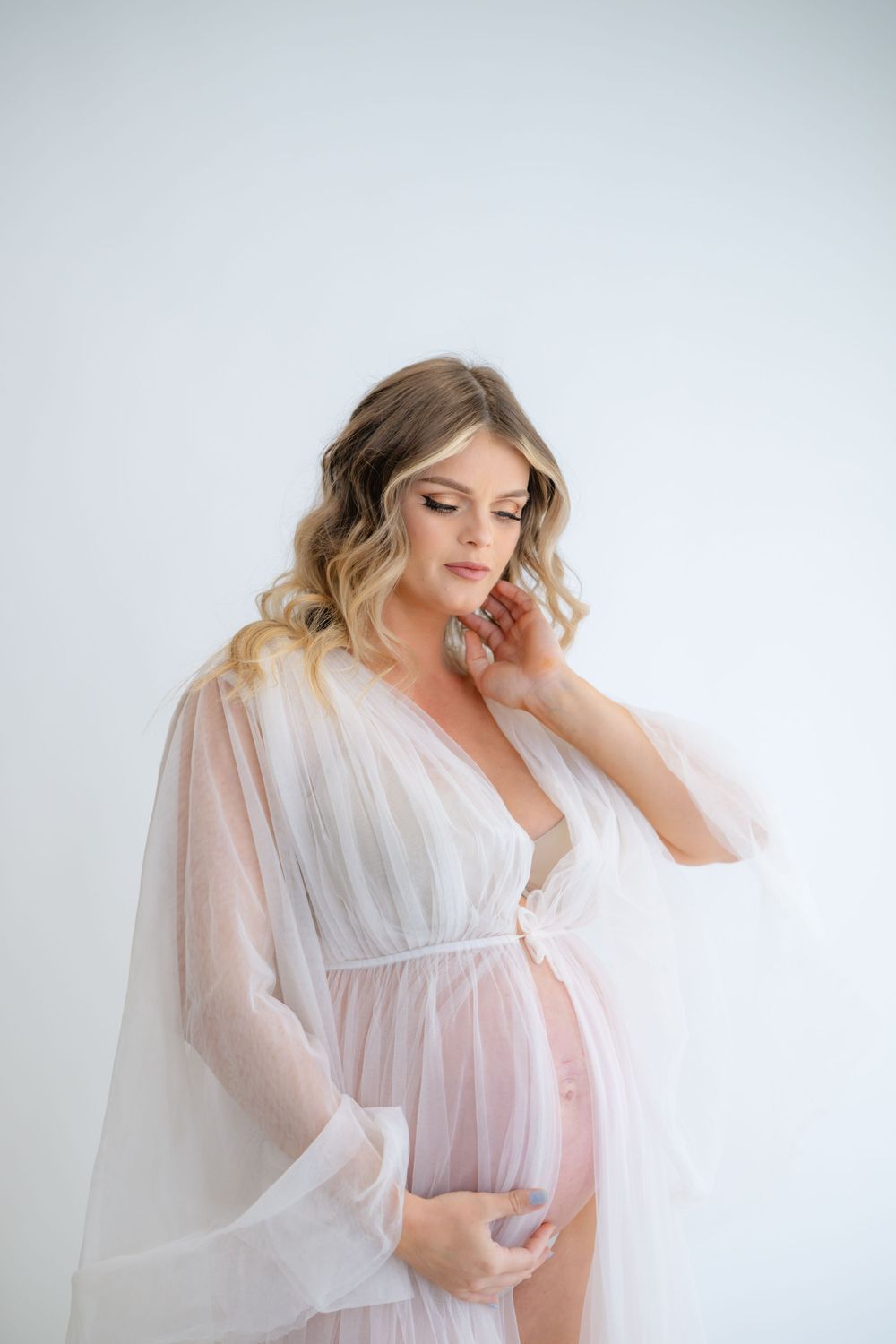 Soft studio maternity portrait in white sheer robe against light background.