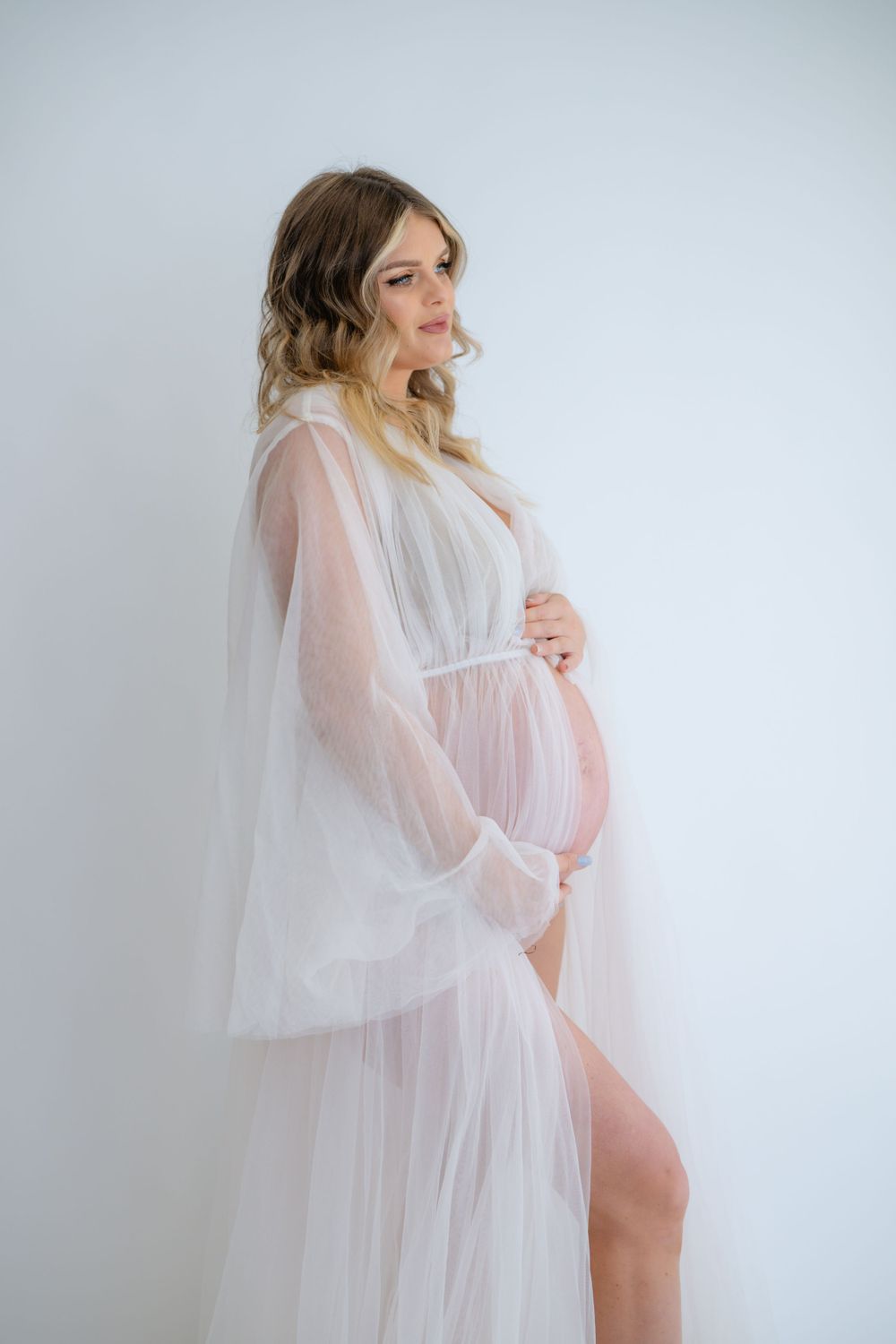 Graceful standing maternity pose in white flowing robe against studio backdrop.