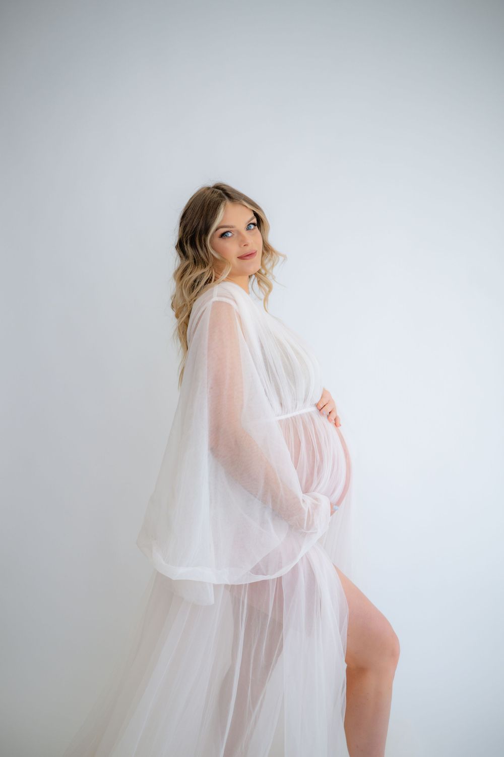 Artistic maternity pose with flowing white fabric in minimalist studio setting.