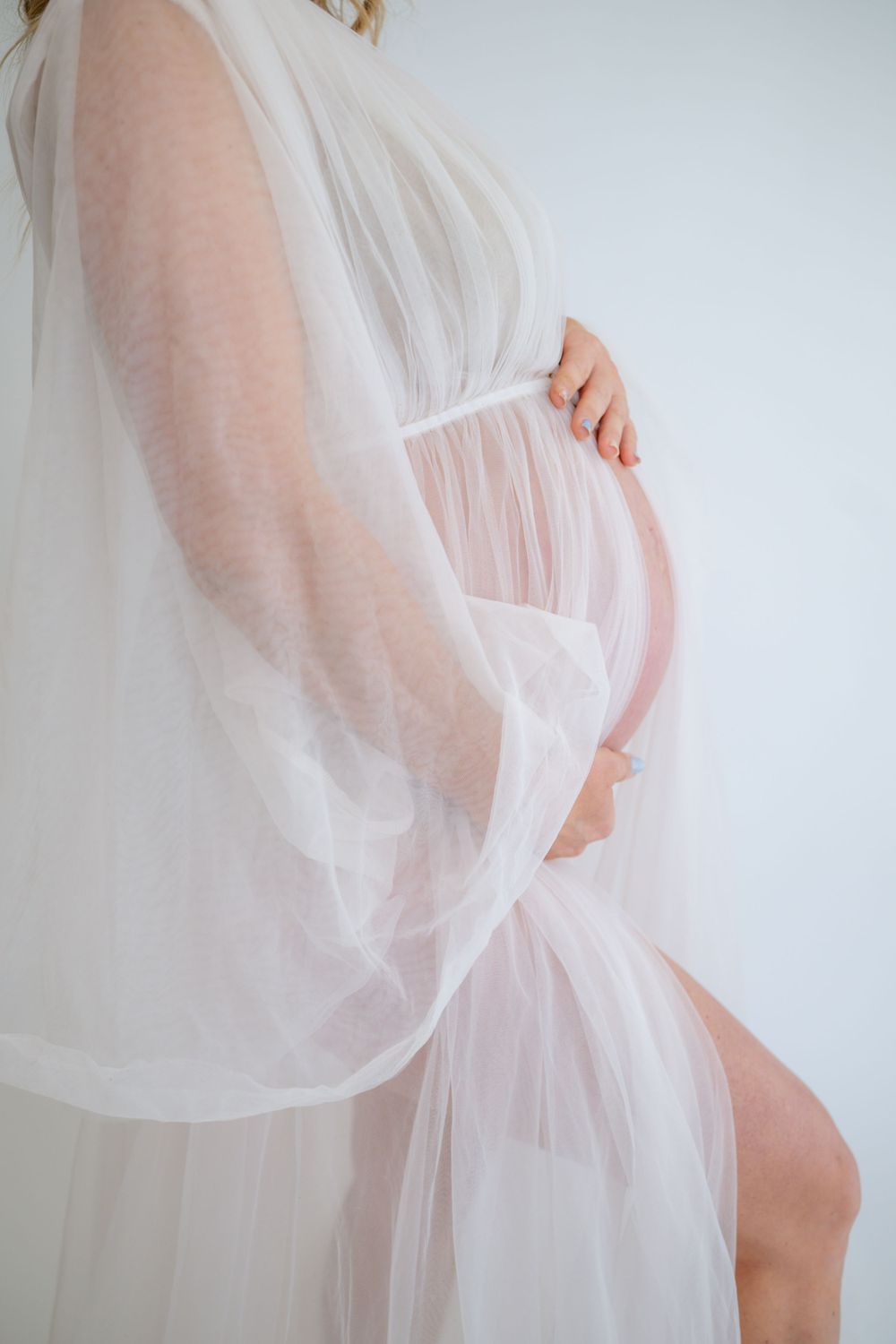 Ethereal white sheer maternity dress drapes elegantly over a pregnant belly in a soft indoor portrait.