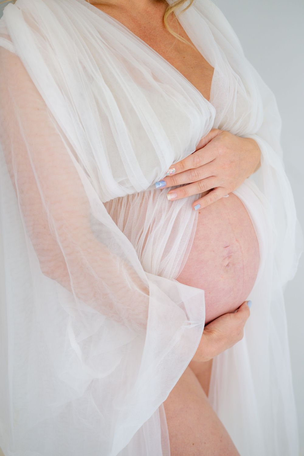 Close-up of pregnant belly draped in delicate white tulle fabric against light background.