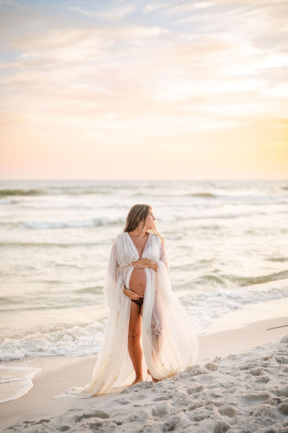 Maternity photoshoot on beach at sunset with flowing white dress and ocean waves.