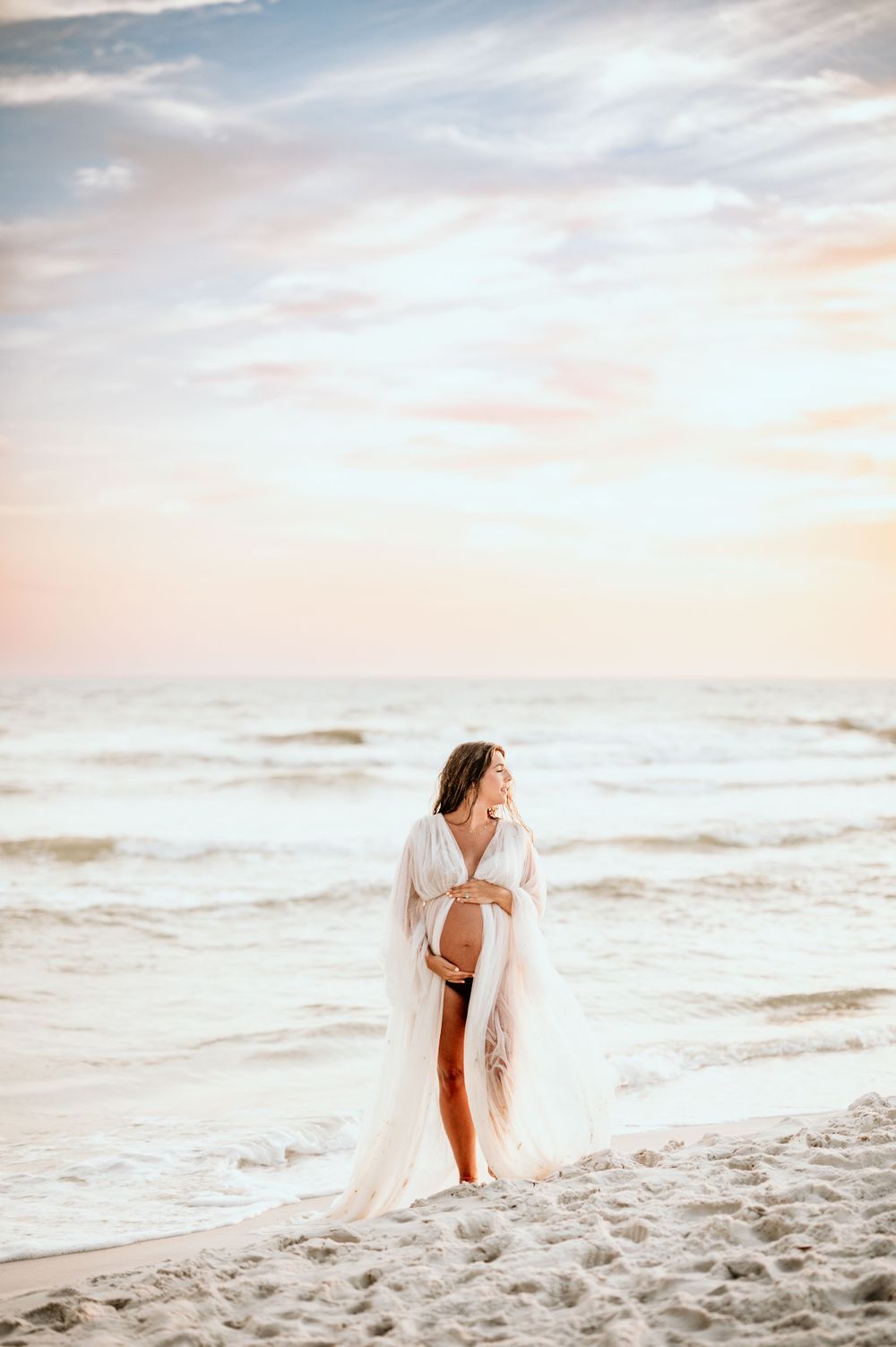 Expectant mother in flowing white dress stands on sandy beach during golden hour.