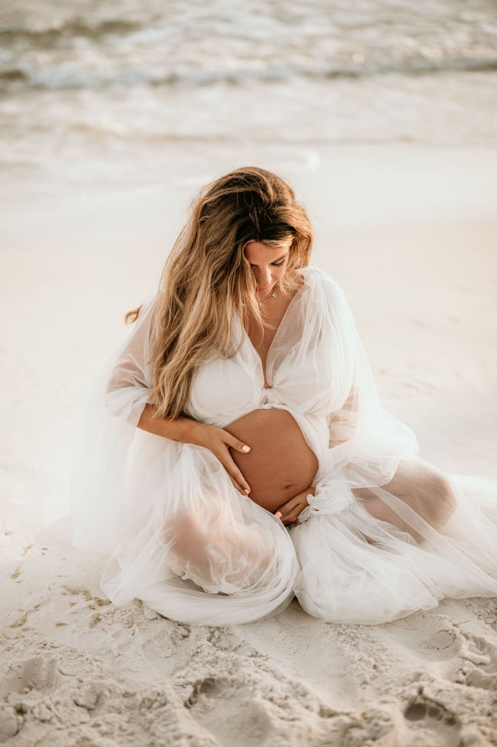 Serene maternity portrait on beach with subject wearing white flowing gown while sitting in sand.