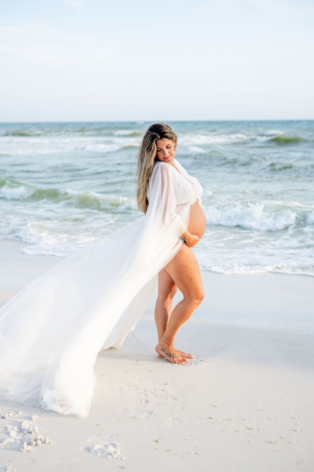Dramatic beachside maternity photo with billowing white dress against ocean waves.