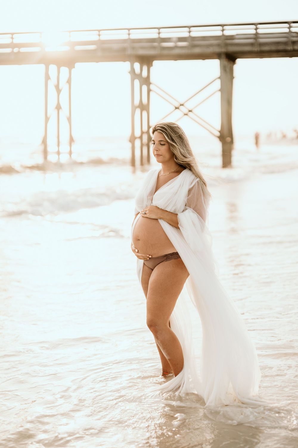 Beachside maternity photo under pier during sunset with flowing white dress.