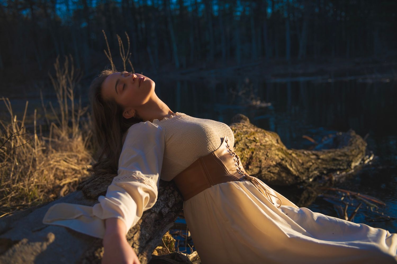 Atmospheric portrait in white dress with brown corset against lakeside setting.