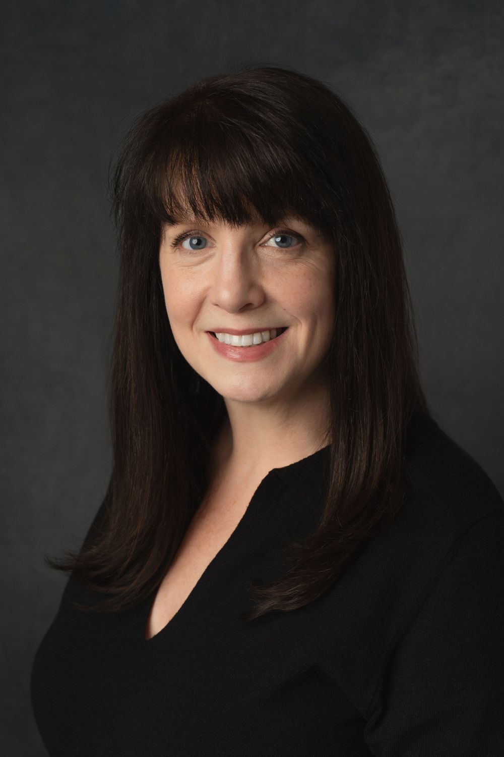 Professional headshot of a smiling woman with dark hair wearing a black top on a gray background.