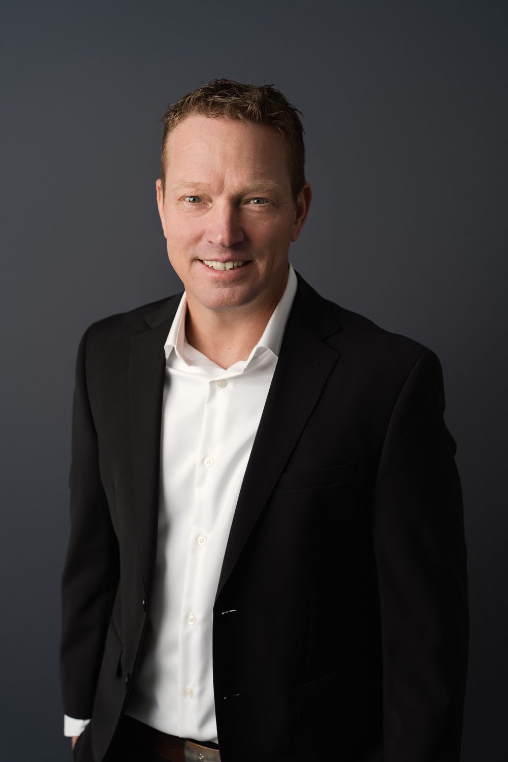 Professional headshot of a smiling man in a black blazer and white shirt against a gray background.