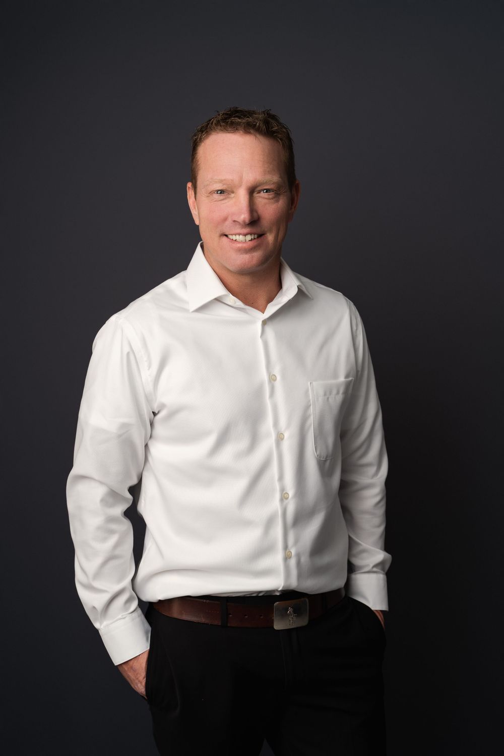 Professional headshot of smiling man in white dress shirt standing against dark gray background.