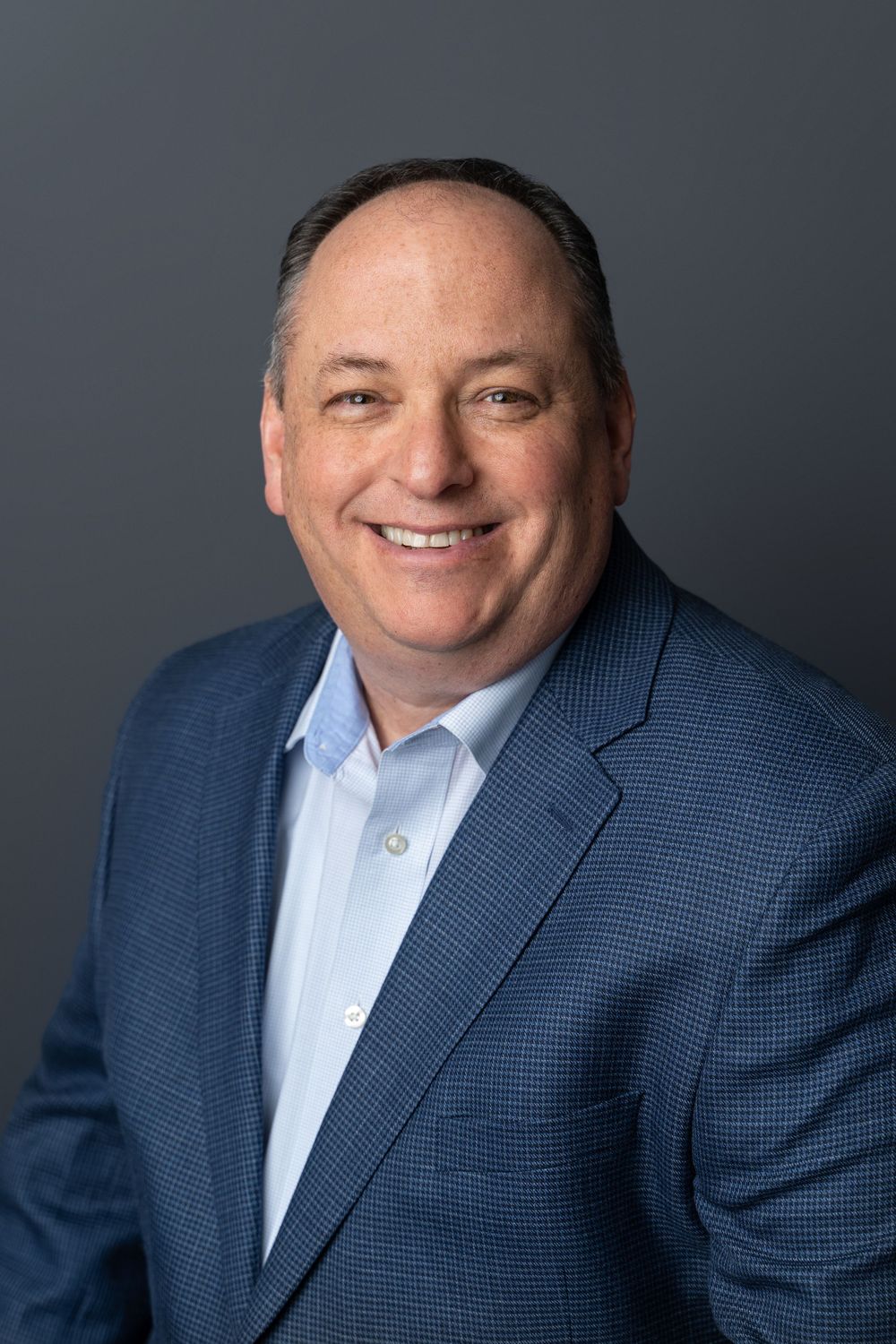 Professional headshot of smiling businessman in blue blazer and light blue shirt on gray background.