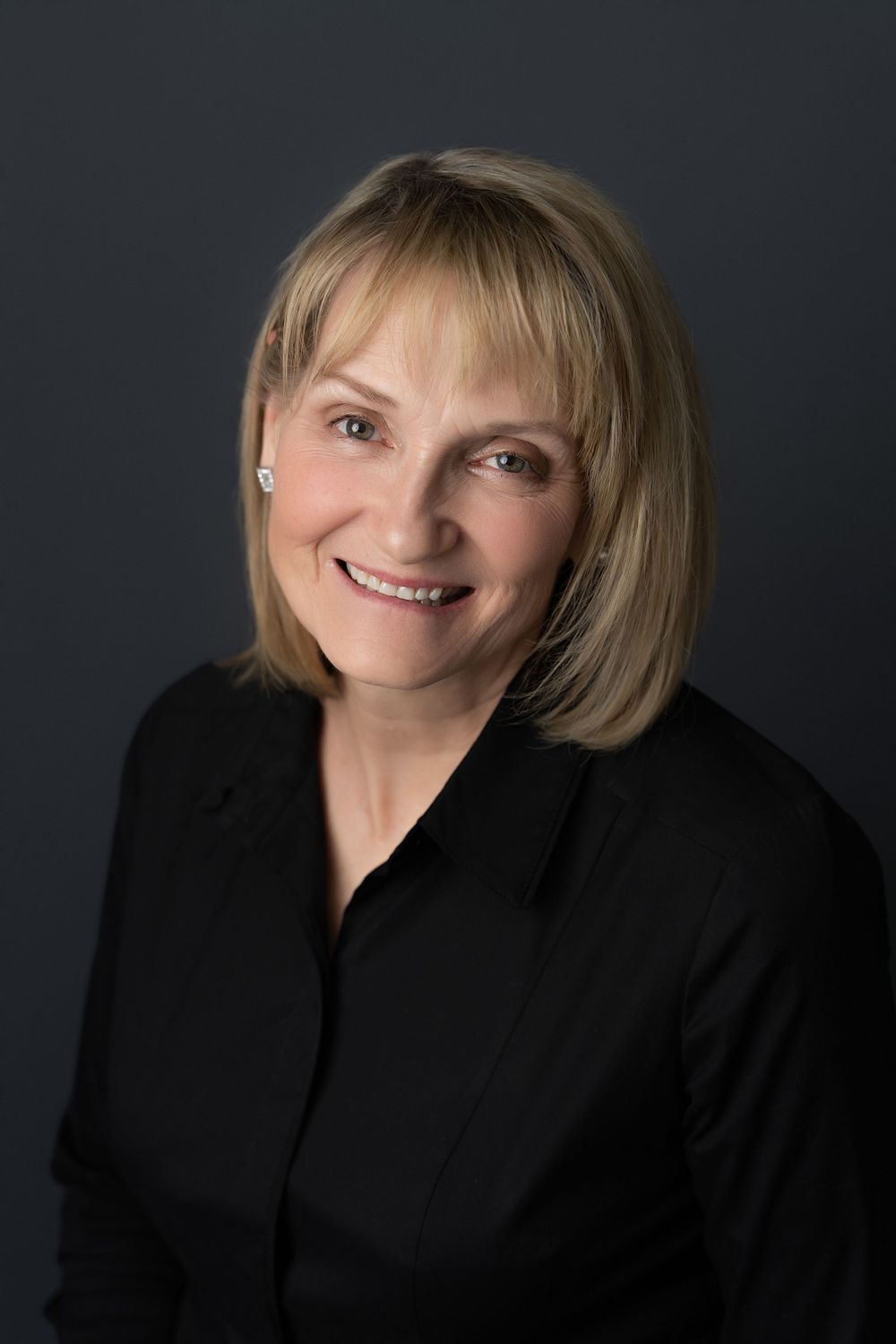 Professional headshot of smiling blonde woman in black blazer against dark gray studio background.