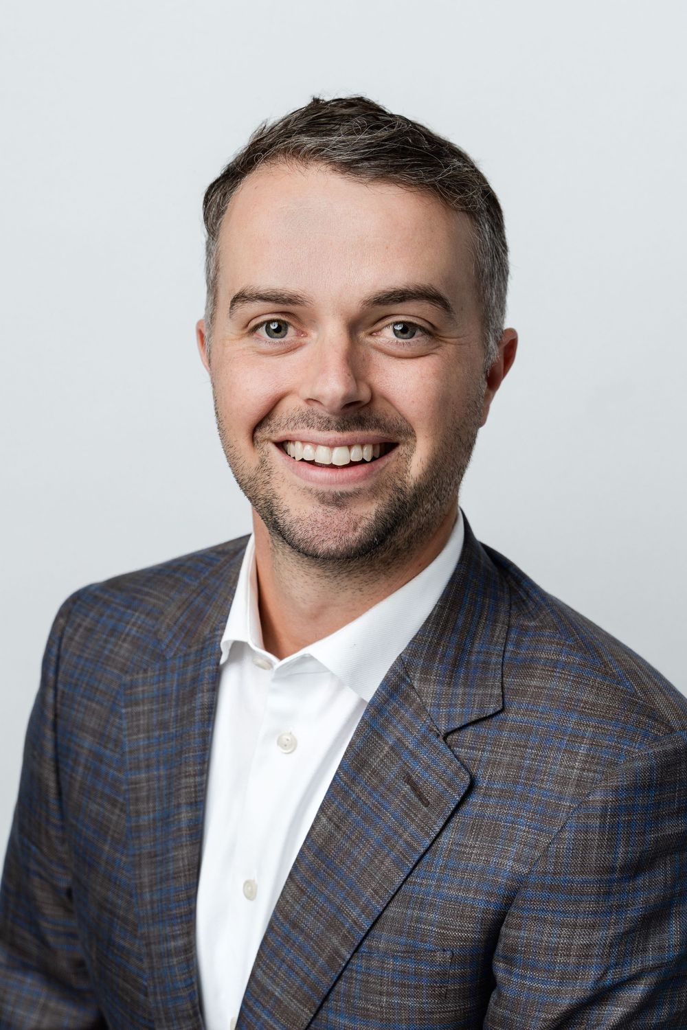 Professional headshot of a smiling man in a plaid blazer and white shirt on a light gray background.
