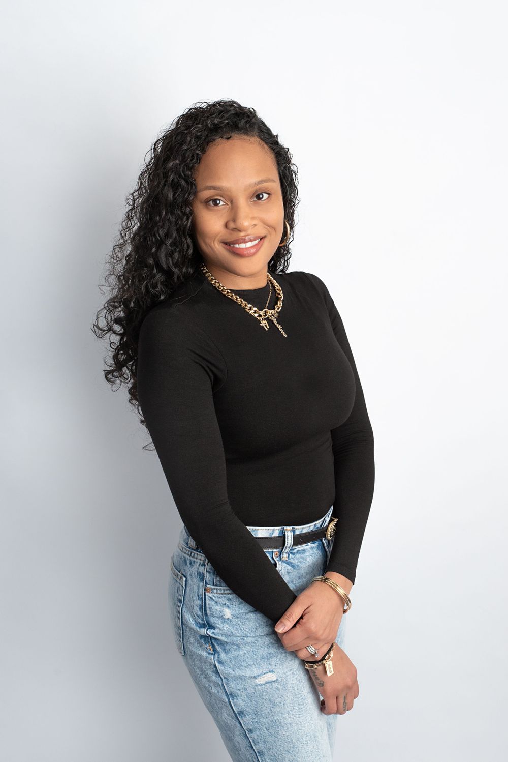 Confident woman with curly hair in a black top and jeans posing against a white background.
