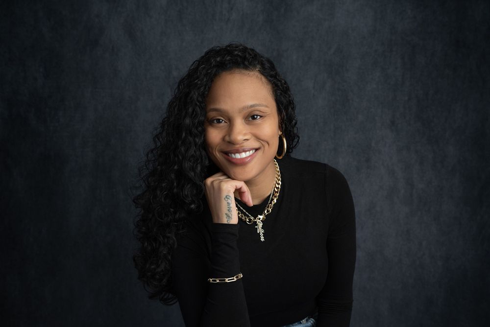 Smiling woman with curly hair wearing a black top and gold jewelry against a dark backdrop.