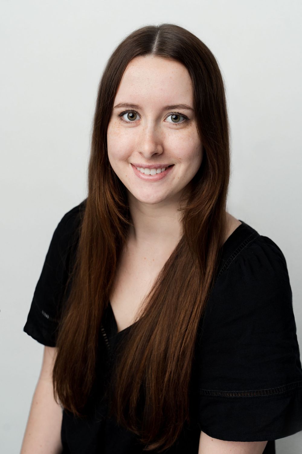 Professional headshot of a young woman with long dark brown hair wearing a black top against a light background.