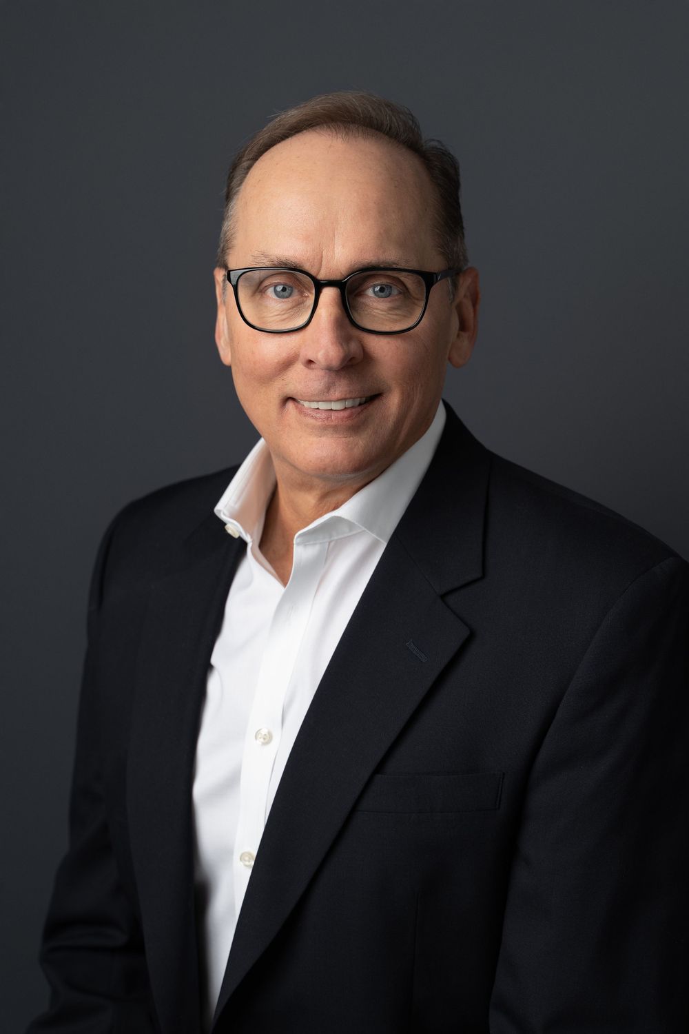 Professional headshot of smiling man wearing glasses and black blazer on dark gray background.