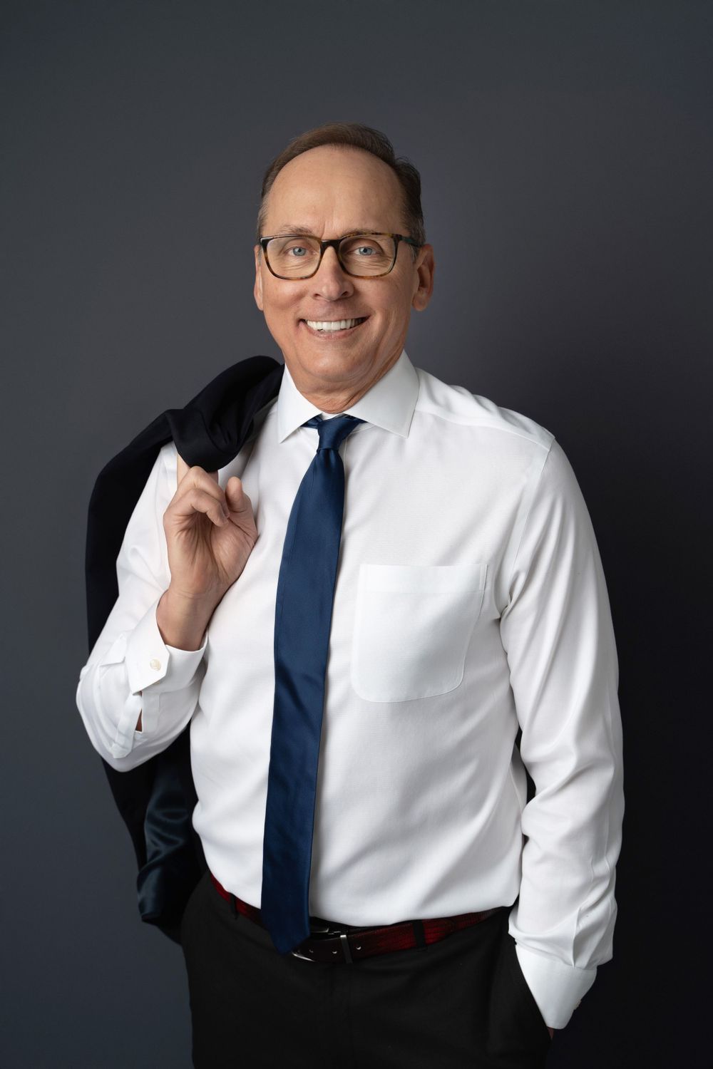 Confident businessman in white shirt and blue tie holding jacket over shoulder on dark background.