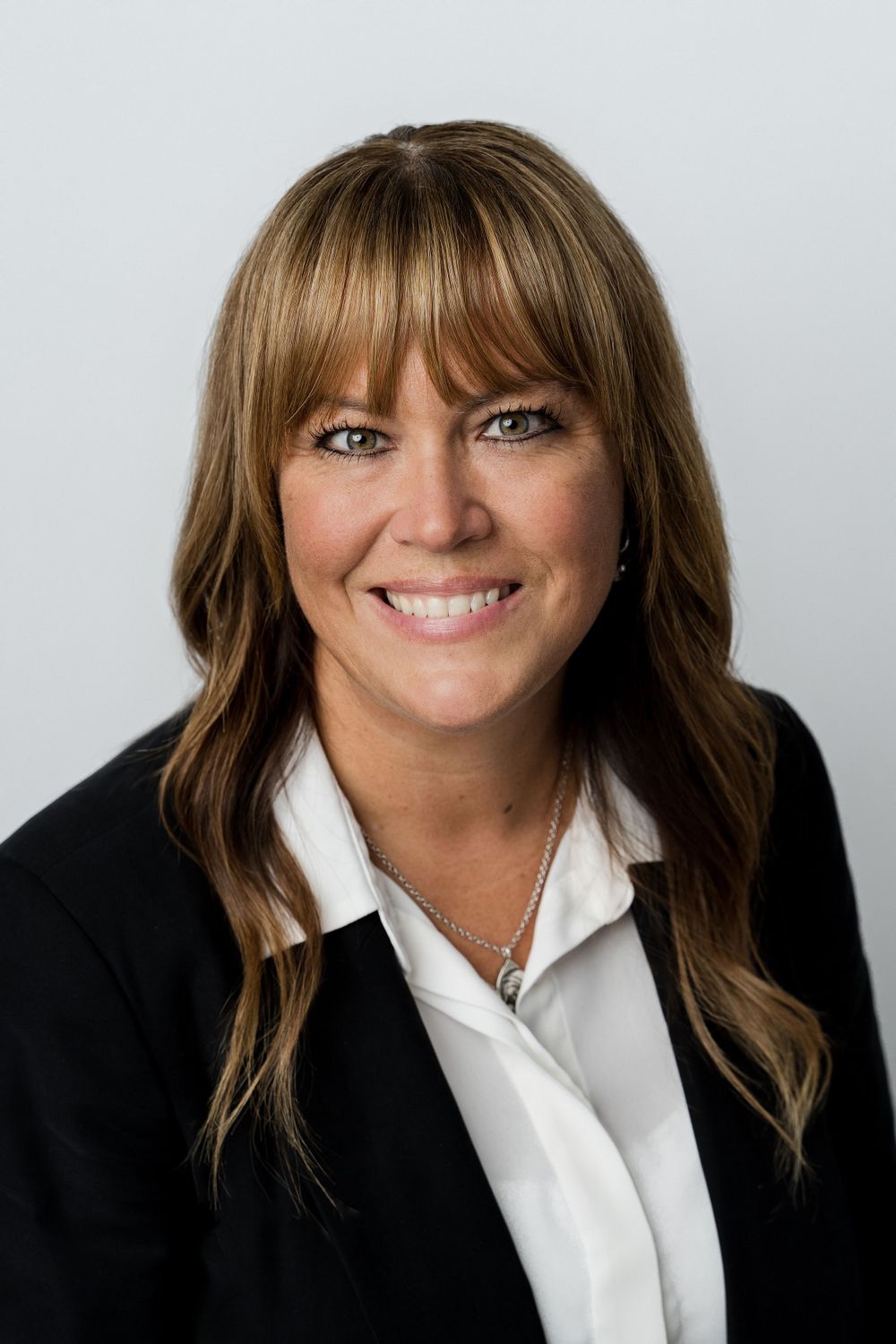 Professional headshot of a smiling blonde woman in a black blazer and white shirt on light background.