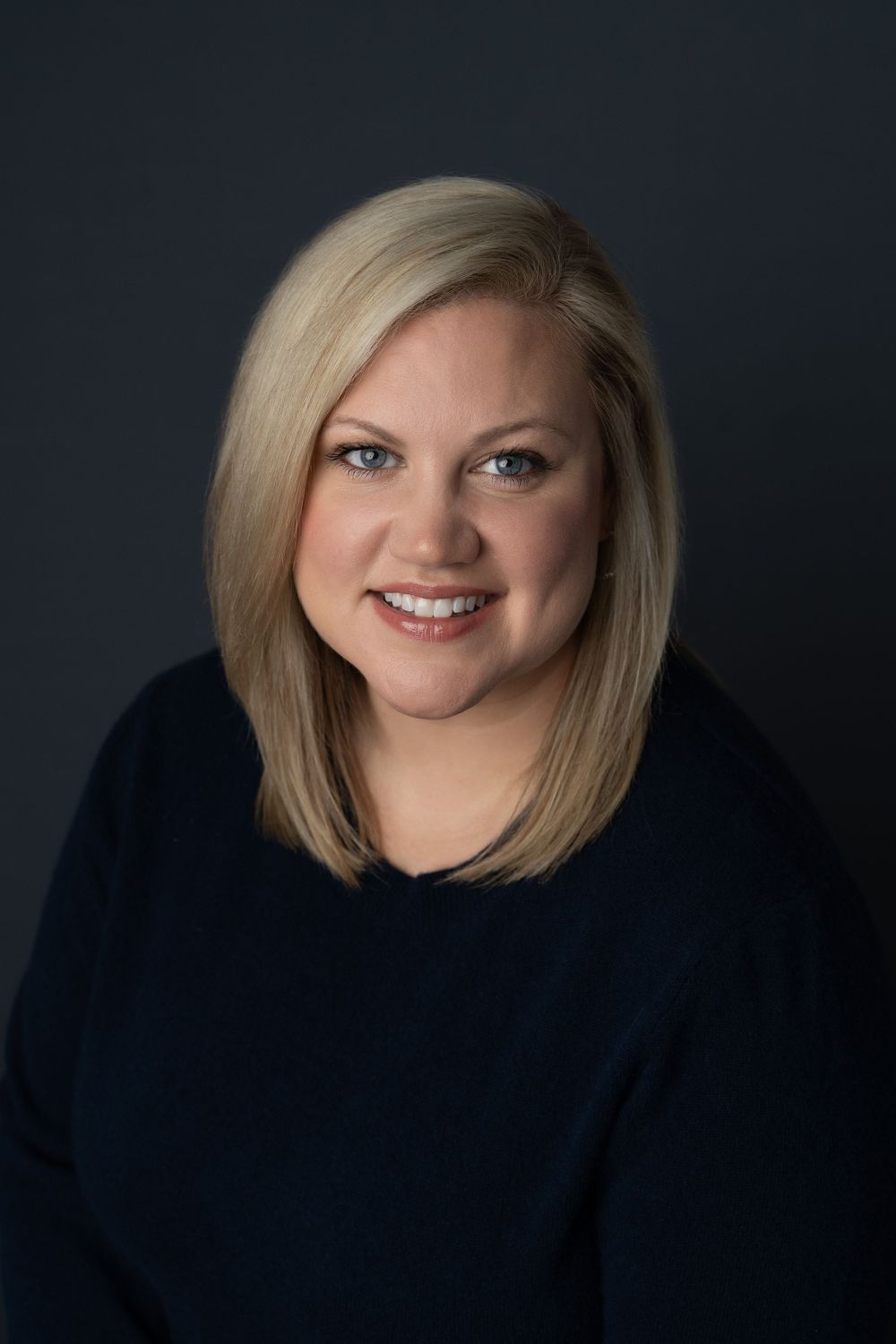 Professional headshot of smiling blonde woman in navy top against dark gray studio background.