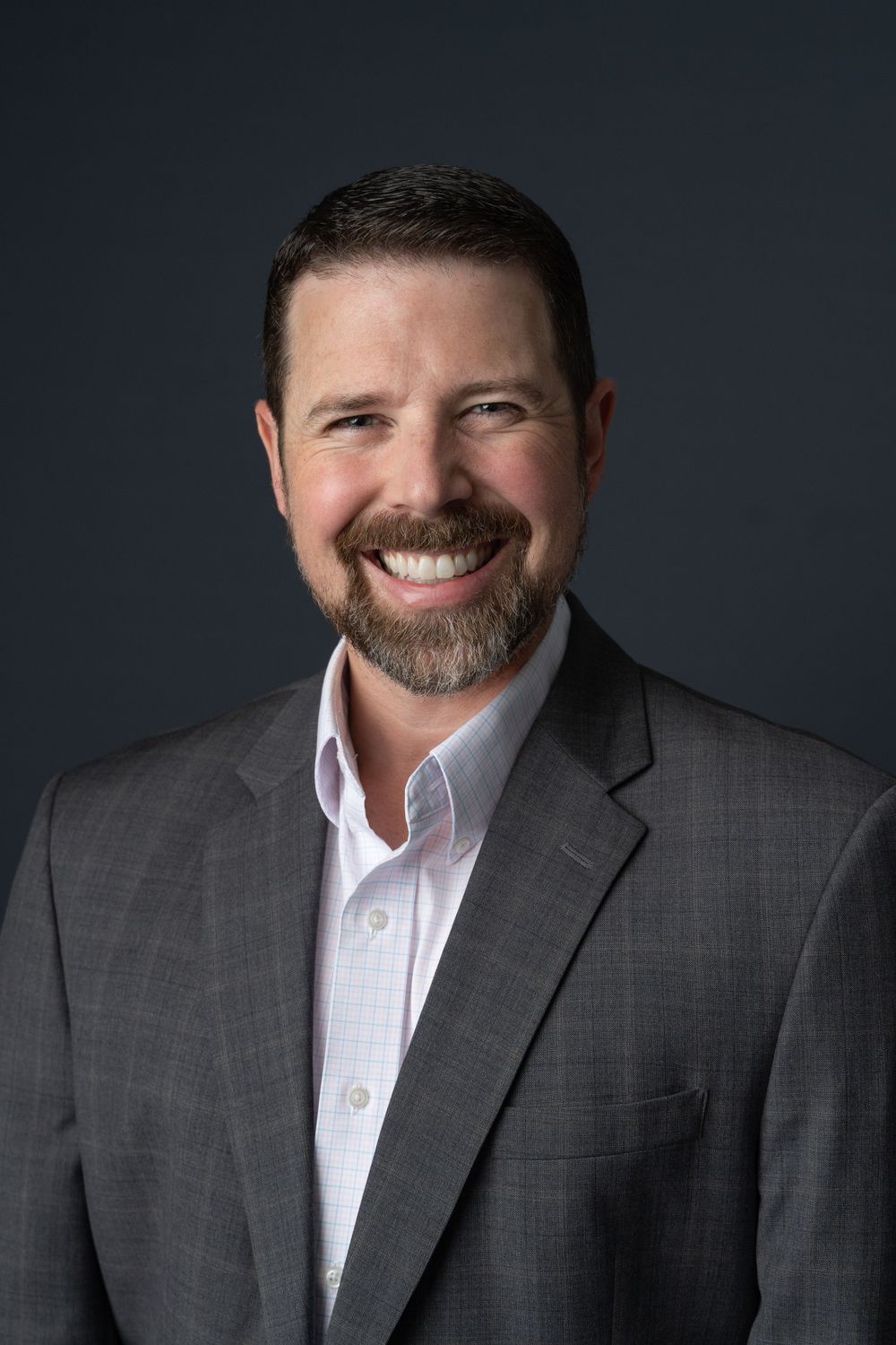 Professional business headshot of smiling man with beard wearing gray blazer on dark background.