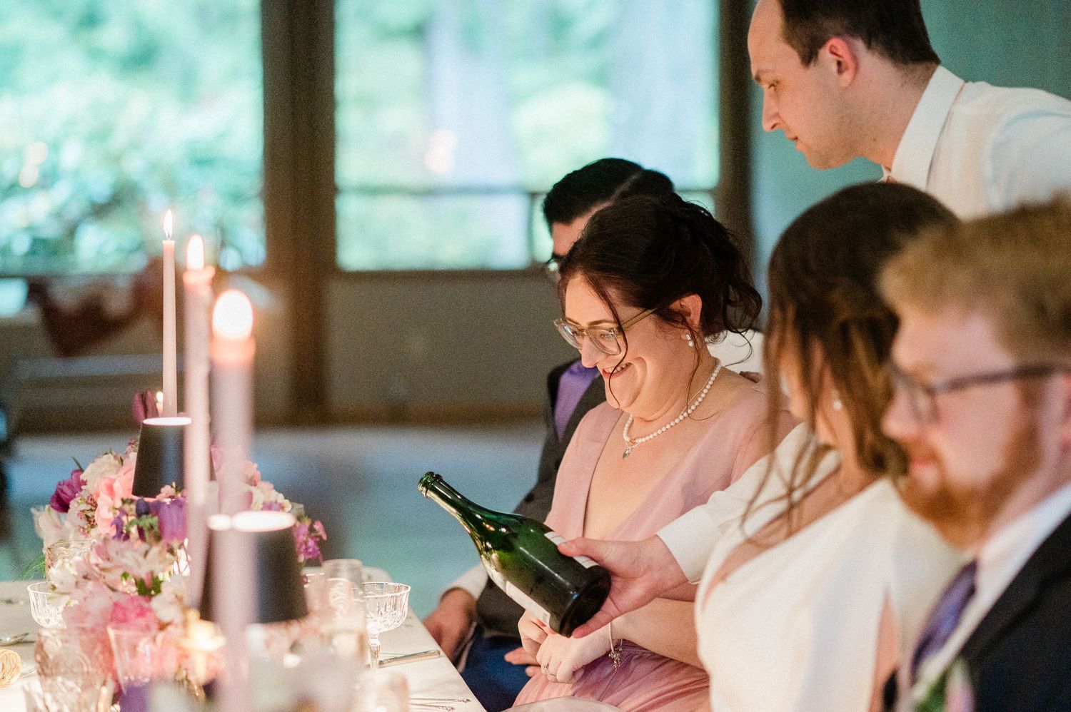 Wedding guests seated at a candlelit dinner table examine a wine bottle during an elegant reception.