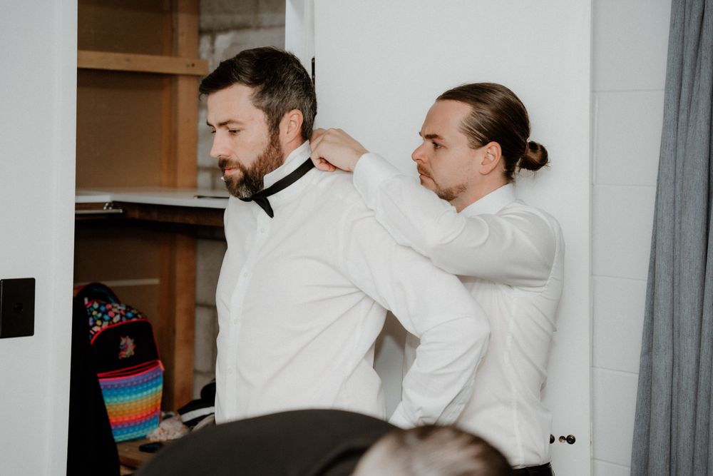 Two people in white dress shirts helping with formal wedding attire preparations.