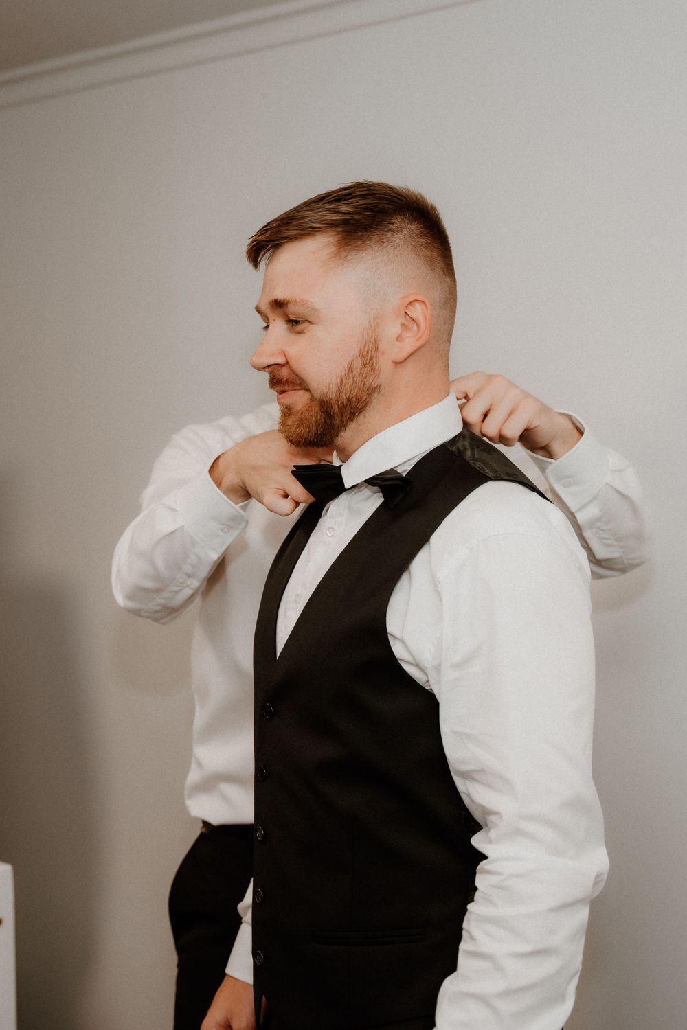 A groom adjusts his bowtie while wearing a formal black vest and white dress shirt.