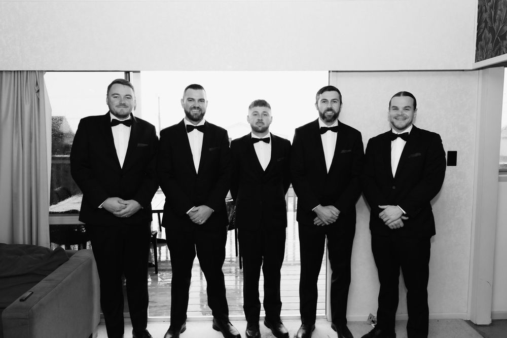 Five groomsmen in matching black tuxedos and bowties stand in formation against a white wall.