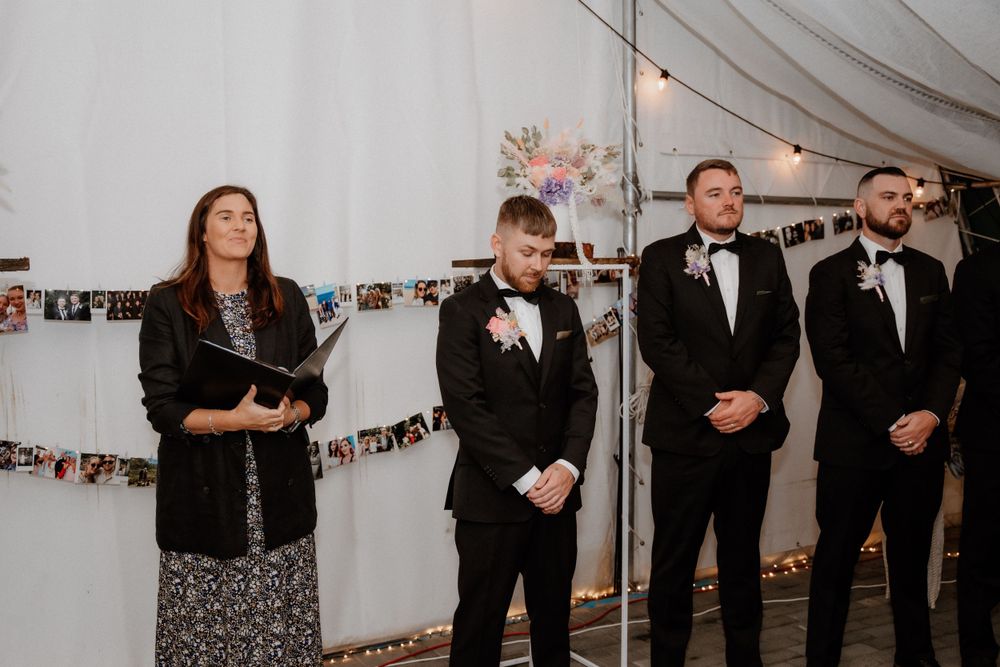 Wedding ceremony setup with officiant and groomsmen standing at altar under draped fabric and photo garland.