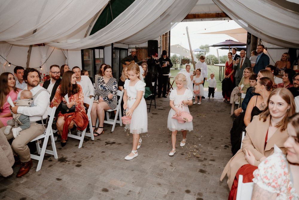 Wedding guests seated in white chairs under tent as flower girls walk down aisle in floral dresses.