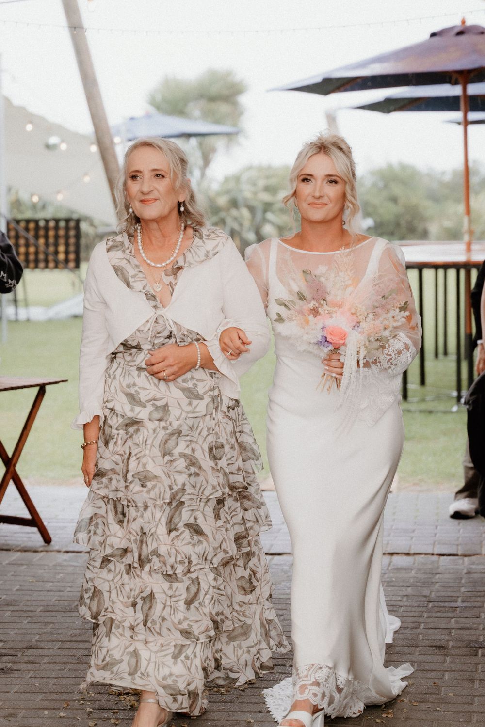 Two women in elegant gowns walk arm-in-arm at outdoor wedding venue.
