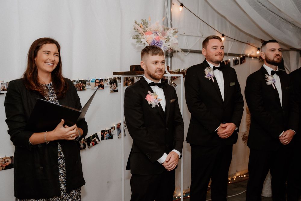 Wedding ceremony scene with officiant and wedding party standing under white draped tent with photo garland.