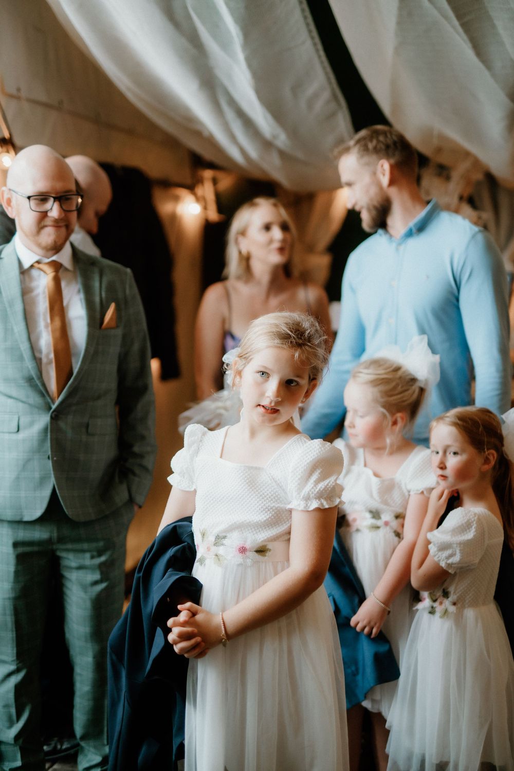 Young attendants in white dresses stand during an indoor wedding ceremony.