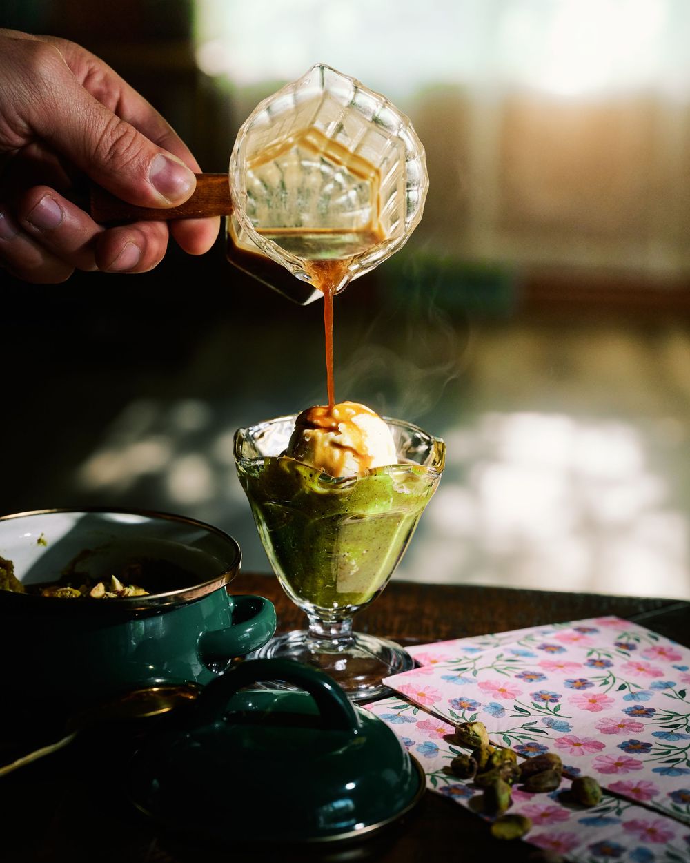 Caramel sauce being poured over a green matcha dessert in a glass with floral placemat and teal dishes.