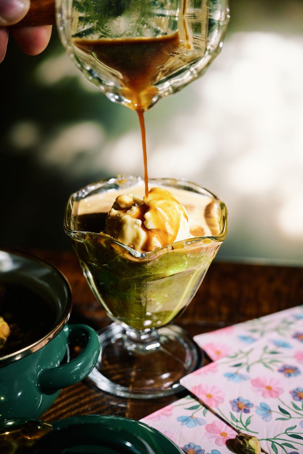 A glass vessel filled with a green dessert being drizzled with caramel sauce over fruit and nuts.