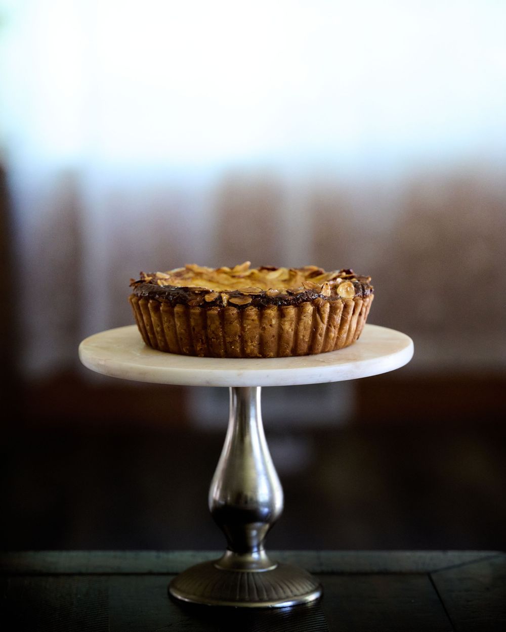 A chocolate tart with chopped nuts sits elegantly on a white pedestal dessert stand against a dark background.