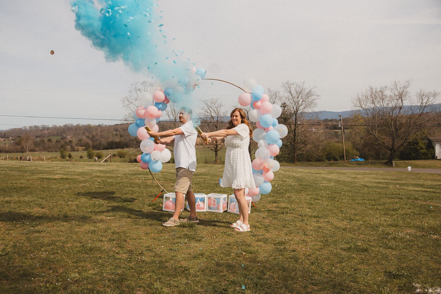 A couple celebrates with blue and pink smoke bombs in a grassy field.