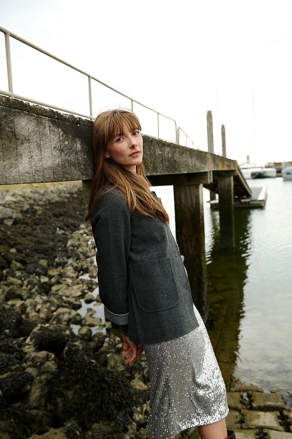 A person in a gray sweater and sequined skirt poses thoughtfully by a waterfront pier on a cloudy day.