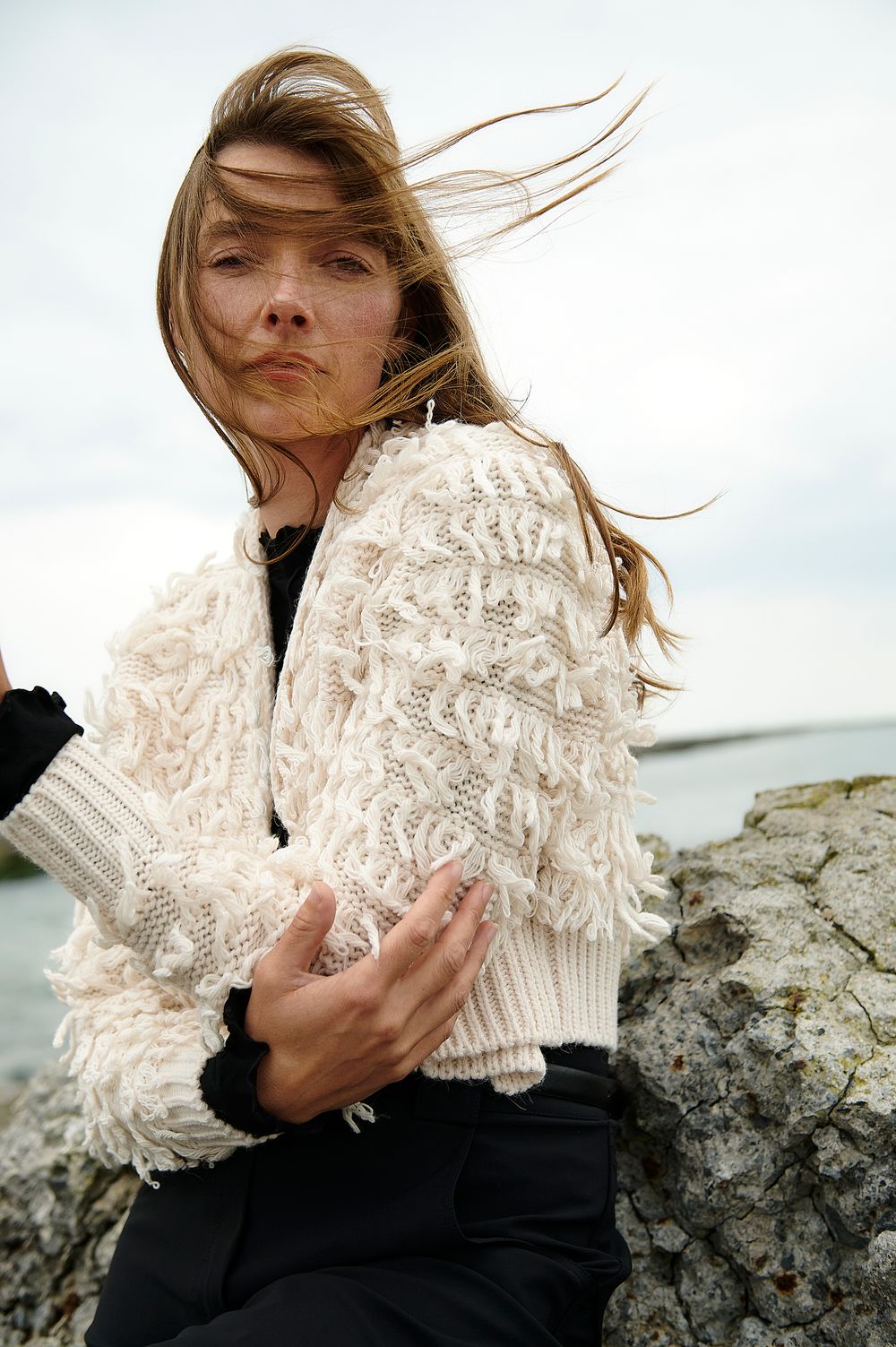 Fashion portrait of a windswept model wearing a textured cream sweater against a cloudy sky by the coast.