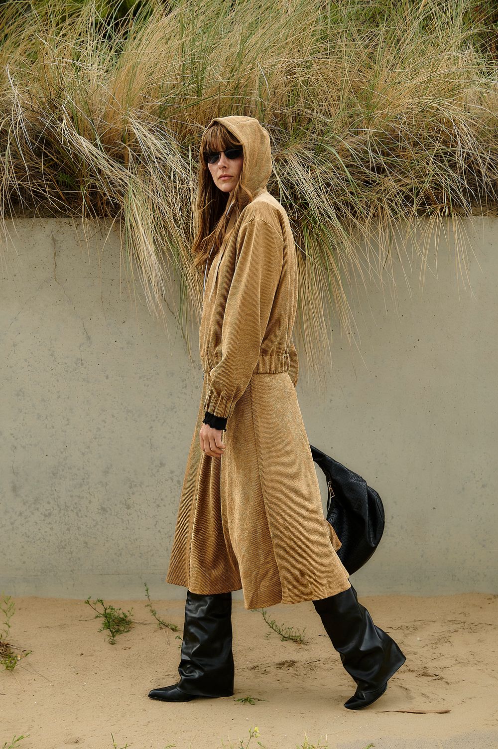 A figure in a tan dress and black boots poses playfully against a sandy beach backdrop with tall grass.