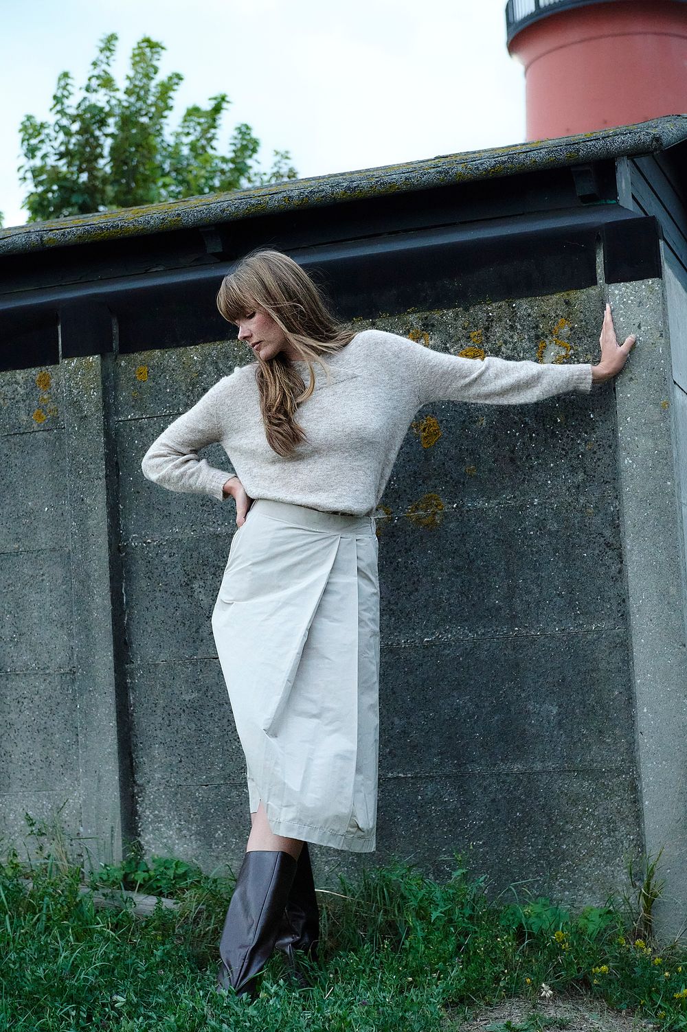 Model in light gray sweater and skirt ensemble poses gracefully against concrete wall near a lighthouse.