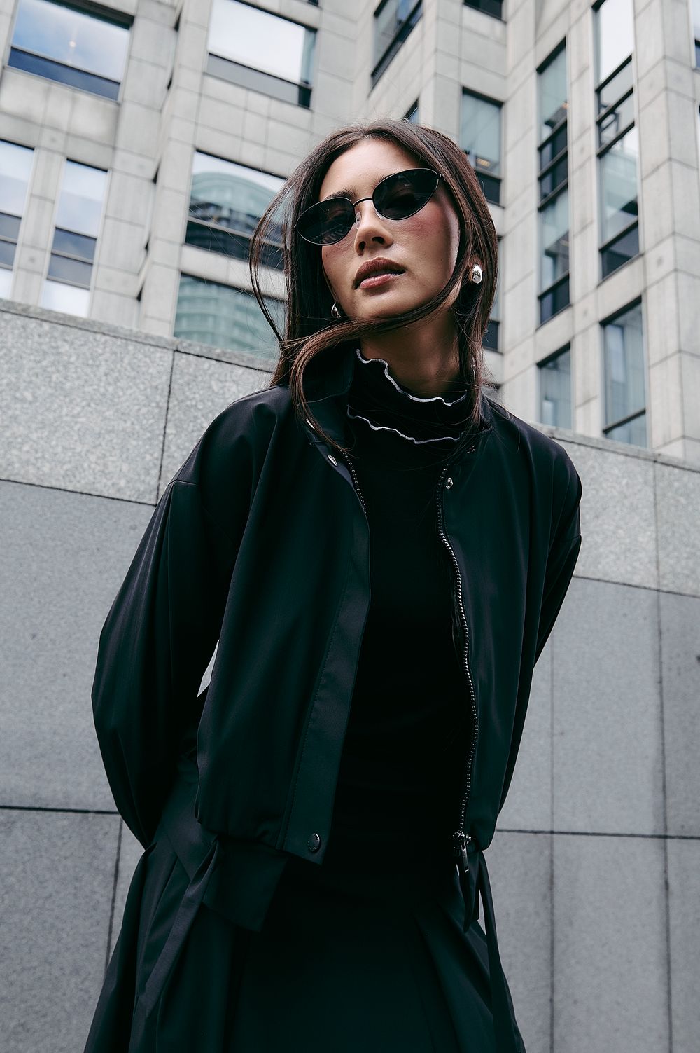 Fashion portrait of a stylish person in sunglasses and black outfit against modern urban architecture.