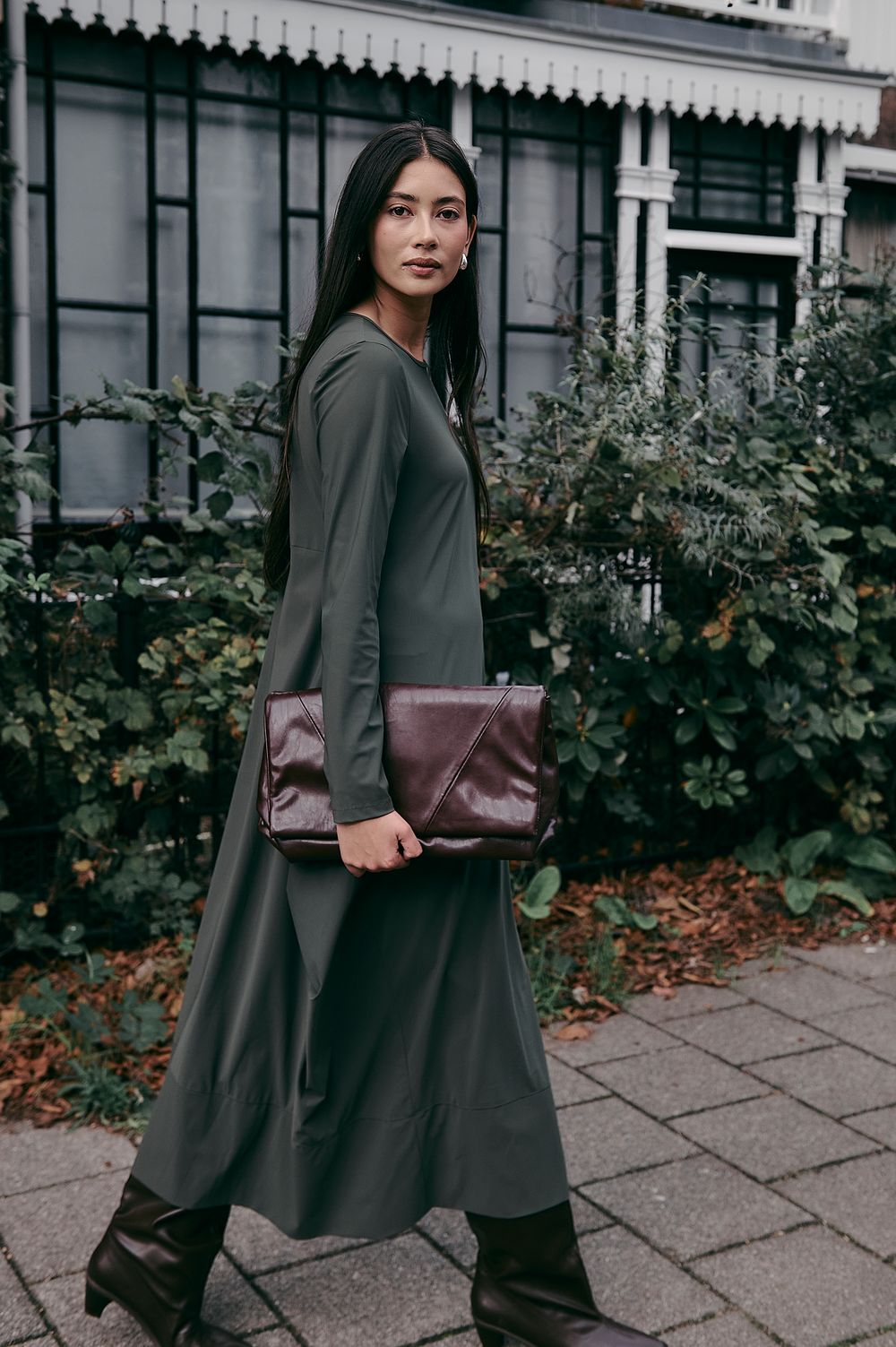 A person in a dark flowing coat carries a burgundy leather clutch while walking along a stone pathway by bushes.